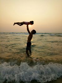 Siblings playing in sea against sky during sunset