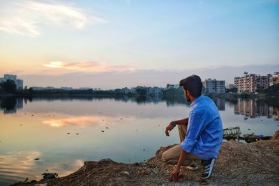 Rear view of man sitting by lake against sky during sunset