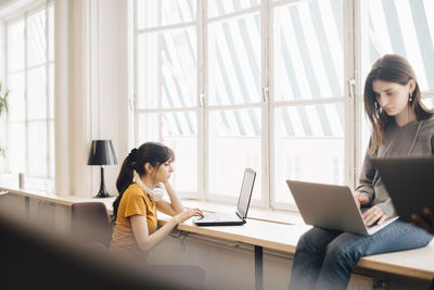 Female colleagues using laptops on desk against window at office