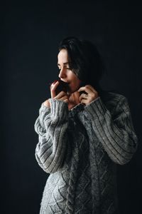 Woman wearing sweater while eating apple against gray background
