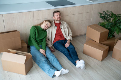 Happy family moving into new apartment, sitting on floor, resting, going to unpack cardboard boxes