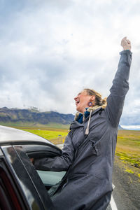 Happy mid adult woman with hand raised screaming while sitting on car window against cloudy sky