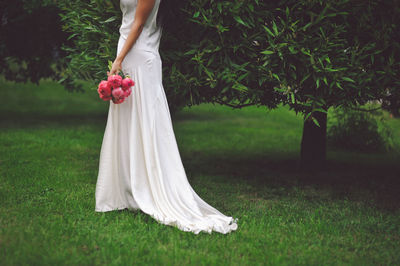 Low section of woman in white dress holding flowers on grassy field