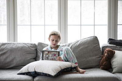 Blonde girl home sick from school plays tablet on couch with teddy