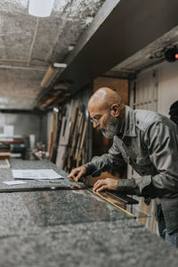 Bald craftsman wearing eyeglasses measuring glass at workbench