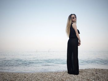 Full length of young woman wearing black dress while standing on shore at beach against sky