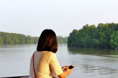 Rear view of woman using phone while standing by lake against clear sky