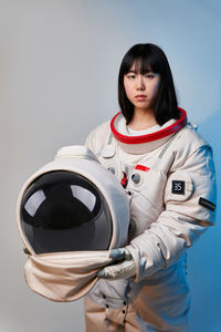 Young asian woman in spacesuit holding helmet and looking at camera under blue light before mission against gray background