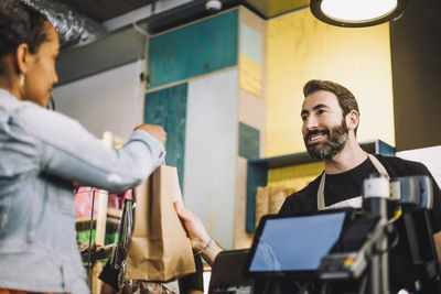 Smiling bearded retail clerk giving shopping bag to female customer at checkout