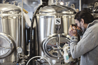 Mature man tasting from container while standing by storage tank in brewery