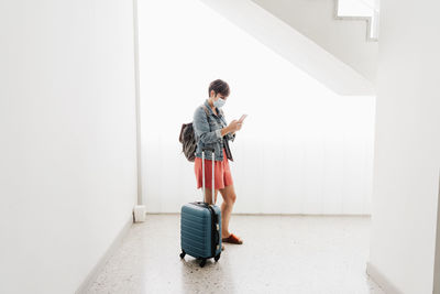 Full length of woman holding mobile phone while standing against wall