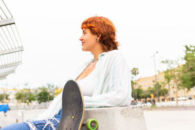 Young female in casual wear with skateboard looking away while sitting in skate park in city