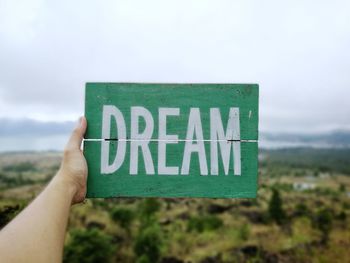 Cropped hand of person holding dream text on wood against sky