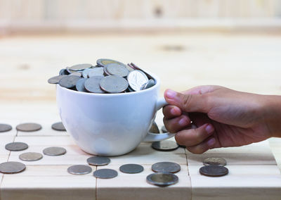 Close-up of hand holding cup with coins on wooden table