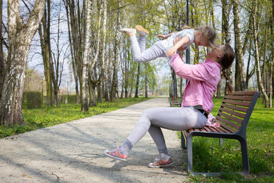 On a bench in a city park, a mother lifts her child high into the air.