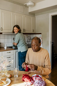 Smiling female nurse looking at senior man knitting in kitchen at home