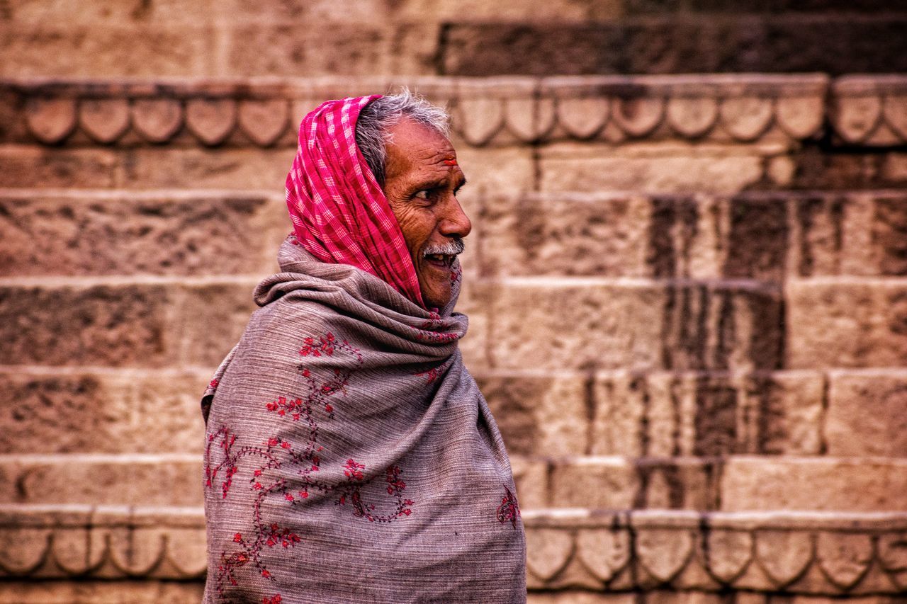 one person, adult, clothing, women, red, architecture, wall, temple, lifestyles, brick wall, brick, headscarf, female, smiling, emotion, standing, traditional clothing, person, looking, scarf, built structure, portrait, wall - building feature, outdoors, happiness, city, focus on foreground, young adult, day, building exterior, casual clothing, waist up, nature