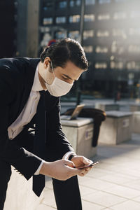 Businessman with face mask sitting outside using phone