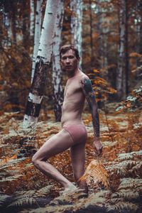 Full length of shirtless man standing by tree in forest