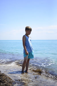 A boy stands in shallow water at sea. he's sad. he looks at the turquoise water and thinks. 