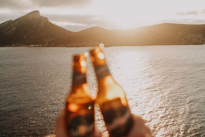 Close-up of hands toasting beer bottle in front of sea