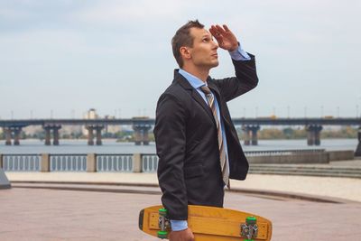 Businessman holding skateboard and saluting while standing outdoors