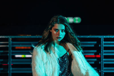 Portrait of young woman standing against illuminated wall at night