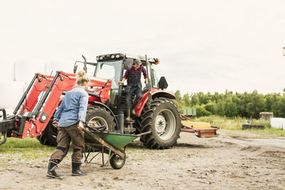 Woman pushing wheelbarrow towards man and tractor at farm