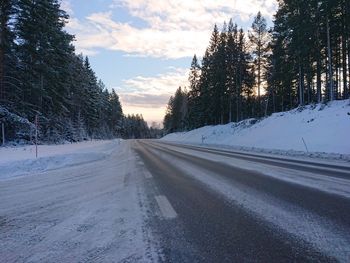 Snow covered road by trees against sky during sunset