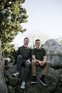 Portrait of happy handsome brothers sitting on rock formations against clear sky at park
