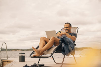 Smiling mature man using laptop while sitting on deck chair at jetty against cloudy sky