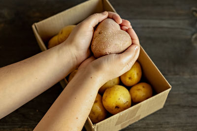 Female hands holding a heart-shaped ugly vegetable potato over a box filled with potatoes. 