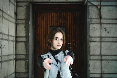 Portrait of young woman pointing towards camera while standing against wall