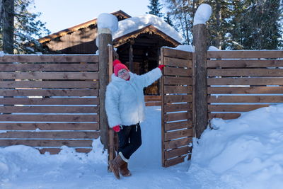 An elderly woman stands near the gate near a wooden house among the snowdrifts in the forest.