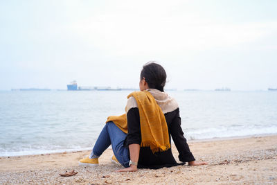 Rear view of woman sitting on shore at beach against sky