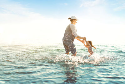 Mother and daughter splashing water in sea against sky