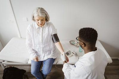 High angle view of male doctor measuring blood pressure of senior patient sitting on bed at medical clinic