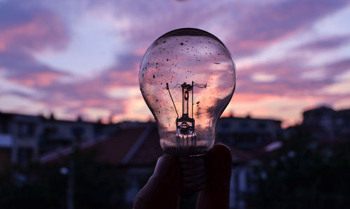 Cropped hand of person holding light bulb against sky during sunset