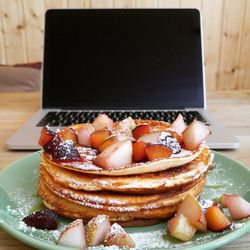 Close-up of pancakes and chopped fruits in plate against laptop on table