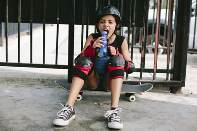 Playful girl drinking water while sitting on skateboard against railings