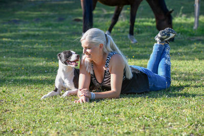 A young blonde woman is lying on lawn next to her black and white dog and looking each others eyes.