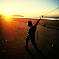 Rear view of silhouette boy with rope standing at beach against sky during sunset