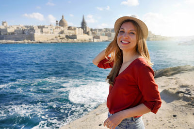 Woman stands on waterfront looking at camera with valletta city on the background, malta island