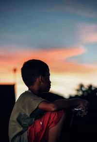 Side view of boy sitting against sky during sunset