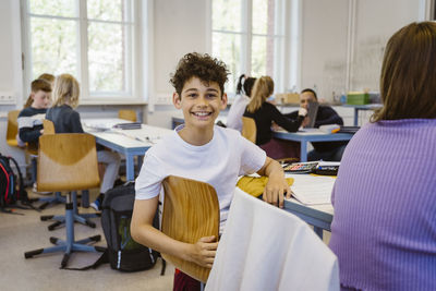 Portrait of happy schoolboy sitting on chair at desk in classroom