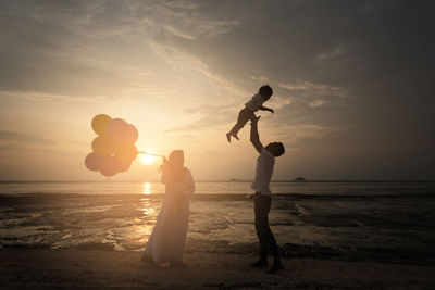 Happy family standing on shore at beach against sky during sunset
