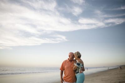 Romantic senior couple looking at each other while standing by sea against sky during sunset