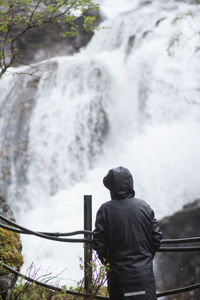 Rear view of teenage girl wearing raincoat standing by waterfall