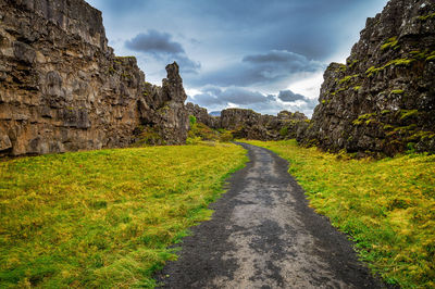 Narrow pathway on grassy field against cloudy sky
