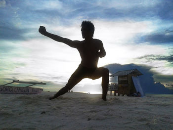 Full length of silhouette young man exercising at beach against cloudy sky during sunset